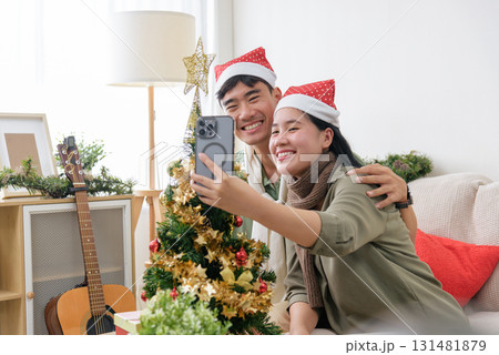 Cheerful couple wearing santa hats takes a selfie together by a decorated tree, they celebrate happy christmas holidays in a cozy home with a guitar visible near the sofa. Cheerful couple wearing santa hats takes a selfie together by a decorated tree, they celebrate happy christmas holidays in a cozy home with a guitar visible near the sofa. 131481879