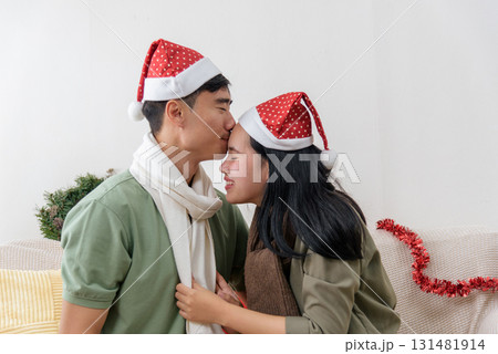 Christmas day brings happy Asian couple love embracing warm winter holiday cheer at home sharing sweet intimate loving moments wearing Santa hats creating special christmas memories for celebration. 131481914