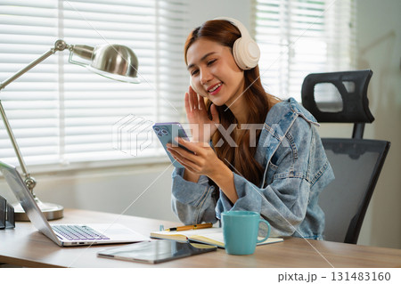 Smiling girl relaxing at home office, she is playing music using smartphone and wearing white headphones. 131483160