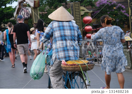 Hoi An Vietnam women in traditional hats sitting on chairs watching tourists and bicycles passing by authentic street life cultural daily scene Asian lifestyle 131488216
