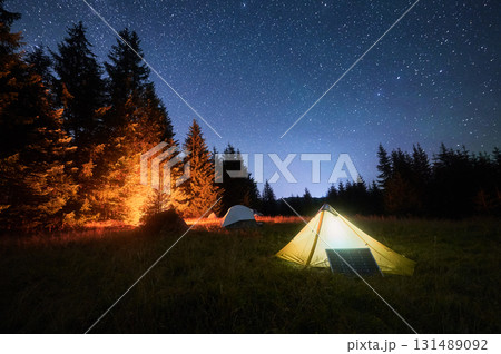 Yellow tent beside solar panel under stunning, star-filled night sky. Nearby, other tents and trees are lit by warm light from campfire, highlighting eco-friendly and serene camping experience. 131489092