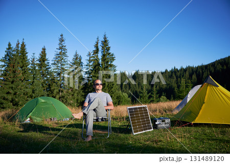 Man using photovoltaic solar panel for charging portable power station outdoor in eco-friendly camping. Male tourist sits relaxed in chair near tents. On background tall pine trees and clear blue sky. 131489120