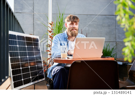 Man works on laptop at outdoor round table, with glass of iced coffee. Solar panel nearby, emphasizing sustainable, modern eco-friendly workspace that combines technology and sustainability. 131489141