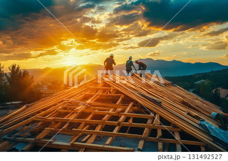 Skilled workers build a wooden roof as the sun sets behind mountains, casting a warm glow and enhancing the scenery above 131492527