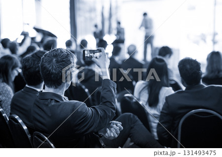 Businessman takes a picture of corporate business presentation at conference hall using smartphone. Business and Entrepreneurship concept. Blue toned grayscale image. 131493475