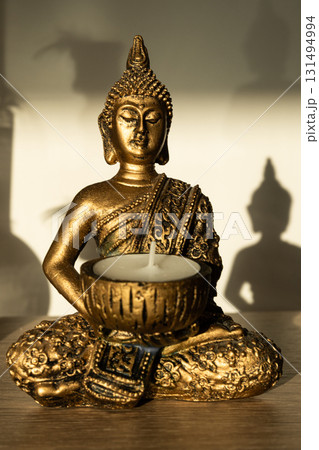 Golden Buddha statue holding a candle holder with a white candle, seated in meditation pose with shadow on beige background. Golden Buddha statue holding a candle holder with a white candle, seated in meditation pose with shadow on beige background. 131494994