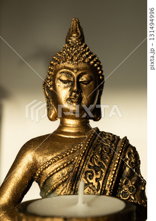 Golden Buddha statue holding a candle holder with a white candle, seated in meditation pose with shadow on beige background. Golden Buddha statue holding a candle holder with a white candle, seated in meditation pose with shadow on beige background. 131494996