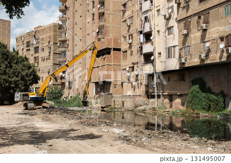 A yellow crane cleans garbage from a polluted lake near residential buildings. The scene shows debris and overgrown vegetation along the water's edge. A yellow crane cleans garbage from a polluted lake near residential buildings. The scene shows debris and overgrown vegetation along the water's edge. 131495007