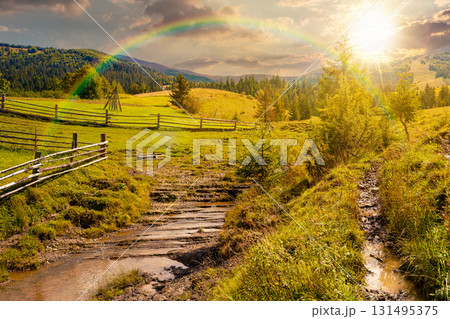 mountainous rural landscape in autumn at sunset. wooden fence near the path through grassy meadow on the hillside. scenery with fir forest on the mountains in evening light under sky with clouds 131495375