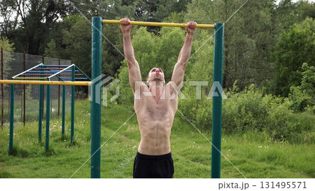 Muscular athlete doing pull ups on horizontal bar at sports ground. Young shirtless sportsman exercising at nature. Strong sporty man training outdoor. Concept of healthy and active lifestyle. Slow mo 131495571