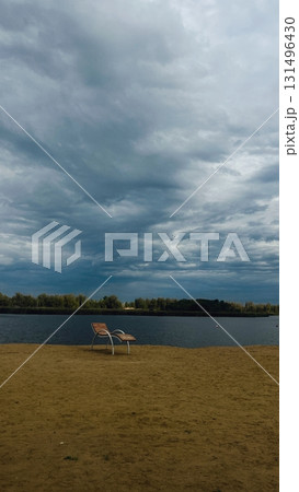 A sun lounger stands on a sandy beach by the river. The beach landscape features a beautiful sky. Dark clouds are in the sky. 131496430