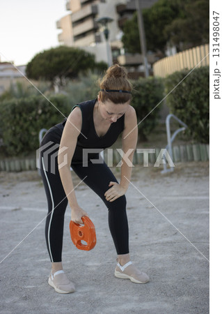 Woman exercising outdoors, holding weight disc, feeling tired 131498047