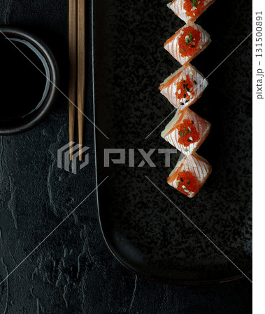 A black rectangular plate showcasing neatly arranged sushi rolls topped with salmon, vibrant red tobiko, and garnished with sesame seeds and herbs.  131500891