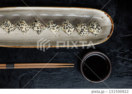 A row of sushi rolls, coated in a mix of black and white sesame seeds, is elegantly arranged on a long ceramic plate.  131500922