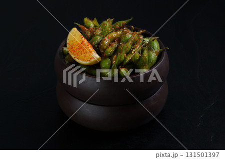 A bowl of seasoned edamame beans garnished with a slice of lemon and sprinkled with chili flakes, creating a vibrant and spicy snack presentation on a dark background. A bowl of seasoned edamame beans garnished with a slice of lemon and sprinkled with chili flakes, creating a vibrant and spicy snack presentation on a dark background. 131501397