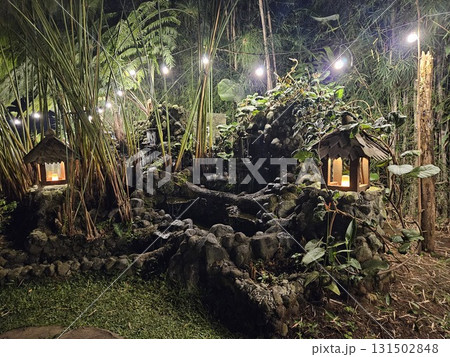 The enchanting nighttime view captures a lush, tropical garden with a small, illuminated hut at its center, surrounded by dense foliage, overhead string lights, and the dark silhouette of trees under  131502848