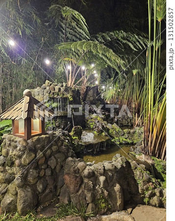 The enchanting nighttime view captures a lush, tropical garden with a small, illuminated hut at its center, surrounded by dense foliage, overhead string lights, and the dark silhouette of trees under  131502857