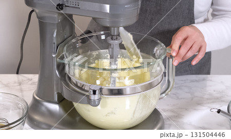 A stand mixer holds creamy cookie dough in a glass bowl set on a marble countertop. The bright light highlights the smooth mixture in a clean kitchen environment. 131504446