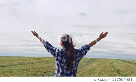 Young woman is embracing the beauty of landscape while raising her arms in the air, feeling the freedom and awe of nature. woman in field with her arms raised up to sky. Young woman is embracing the beauty of landscape while raising her arms in the air, feeling the freedom and awe of nature. woman in field with her arms raised up to sky. 131504481