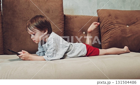 A three-year-old preschooler is lying on the couch, playing with his phone and actively scrolling through social media. boy playing video games on his phone on sofa. Video game addiction A three-year-old preschooler is lying on the couch, playing with his phone and actively scrolling through social media. boy playing video games on his phone on sofa. Video game addiction 131504486
