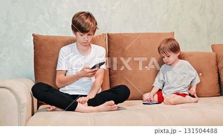 three-year-old preschooler and teenage boy are sitting on the couch, playing with their phones, actively browsing social media. boys are playing video games on phones on couch. Video game addiction 131504488
