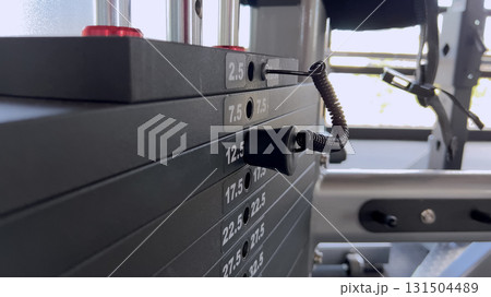 Detailed view of a fitness machine showing numbered weight plates and a cable. The equipment is designed for resistance-based strength training. 131504489
