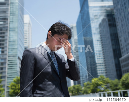 頭を抱えるスーツ姿の日本人男性がビル群の前で落ち込む仕事のストレスやビジネスの悩み 131504670