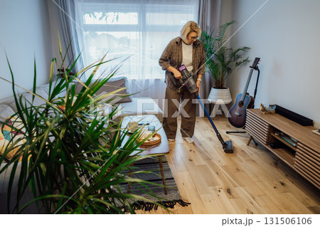 Middle aged woman is vacuuming floor of modern apartment living room with green plants, using portable cordless vacuum cleaner. Daily routine and household chores. Housekeeping, home cleaning concept. Middle aged woman is vacuuming floor of modern apartment living room with green plants, using portable cordless vacuum cleaner. Daily routine and household chores. Housekeeping, home cleaning concept. 131506106