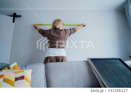 Back view Middle aged woman doing measuring with a measure tape on the wall. Lady wants to put a picture on the wall at home. Housekeeping work. Doing repair herself. DIY, equality in work concept. 131506127