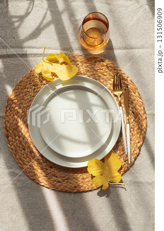 Table Setting with Yellow Leaves on White Plate and Woven Placemat under Sun Light 131506909