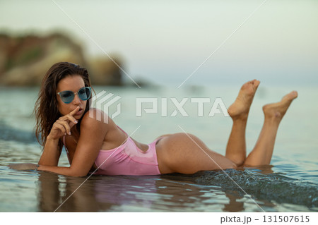 beautiful woman lies on her stomach in shallow water at the beach. The sunset reflects on the calm sea as she wears sunglasses and playfully gestures for silence. beautiful woman lies on her stomach in shallow water at the beach. The sunset reflects on the calm sea as she wears sunglasses and playfully gestures for silence. 131507615