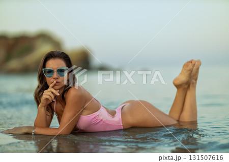 woman in a pink swimsuit lies on her stomach in shallow water playfully putting her finger to her lips as the sun sets in the background. Calm waves gently lap against her. woman in a pink swimsuit lies on her stomach in shallow water playfully putting her finger to her lips as the sun sets in the background. Calm waves gently lap against her. 131507616