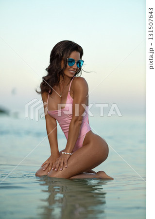 beautiful woman kneels in shallow water at the beach enjoying the ocean waves and the warm sunset. She wears a pink swimsuit and stylish sunglasses smiling and soaking in nature. 131507643