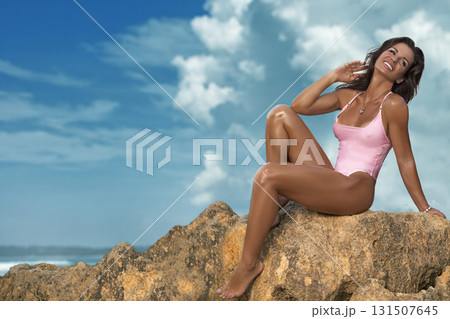 beautiful woman sits on a large rock by the seashore wearing a pink swimsuit. She smiles happily while enjoying the sunny weather and the refreshing ocean breeze. beautiful woman sits on a large rock by the seashore wearing a pink swimsuit. She smiles happily while enjoying the sunny weather and the refreshing ocean breeze. 131507645