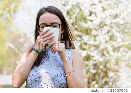 Outdoor shot of discontent young girl with seasonal allergy, uses white tissue, poses over blooming tree, has rhinitis and sneezing, reacts on allergens. People and disease concept 131507698