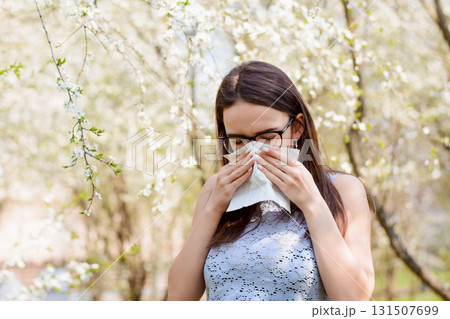 Young student girl hiding her face with white light handkerchief while sneezing. Caucasian female with running nose blows her nose against spring trees. Concept of spring Illness Young student girl hiding her face with white light handkerchief while sneezing. Caucasian female with running nose blows her nose against spring trees. Concept of spring Illness 131507699