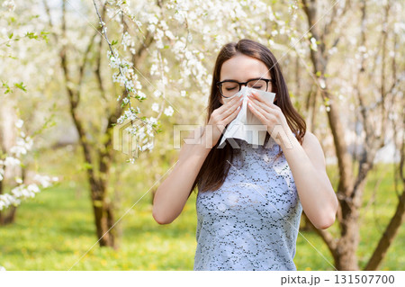 Portrait of an allergic young girl in the park holding white napkin and sneezing because of allergy on pollen of blooming trees Portrait of an allergic young girl in the park holding white napkin and sneezing because of allergy on pollen of blooming trees 131507700