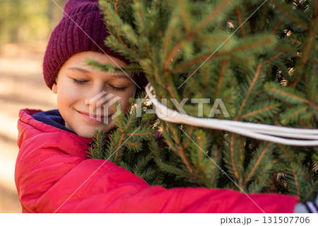 Cute boy in a warm red hat and jacket hugging a christmas tree. Happy kid with closed eyes and a smile on his face can't wait for decorating a tree for winter holidays. Cute boy in a warm red hat and jacket hugging a christmas tree. Happy kid with closed eyes and a smile on his face can't wait for decorating a tree for winter holidays. 131507706