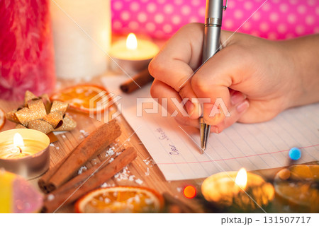 Close-up shot of hand of a child, writing a letter to Santa. Christmas decoration around, festive atmosphere, belief in magic and miracle. Close-up shot of hand of a child, writing a letter to Santa. Christmas decoration around, festive atmosphere, belief in magic and miracle. 131507717
