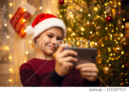 Cute young boy makes a selfie in the hat of Santa Claus, decorated fir-tree full of lights on the background. The concept of Christmas, holidays. Cute young boy makes a selfie in the hat of Santa Claus, decorated fir-tree full of lights on the background. The concept of Christmas, holidays. 131507723
