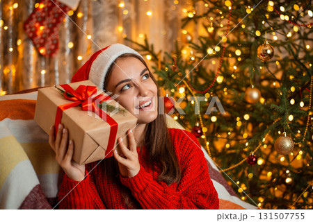 Young pretty girl in a good mood trying to guess what the gift box contains. Christmas tree and warm yellow light of garlands on the background, festive atmosphere. 131507755