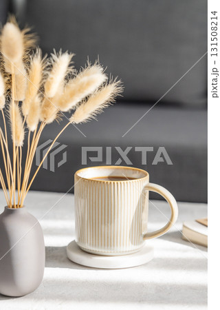 Beige cup of coffee on a light table with soft shadows, vase and notebook on the background 131508214