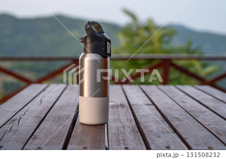 Stainless steel water bottle with black lid on rustic wooden table with nature and mountains  131508232