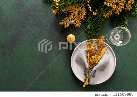 Top view of a Christmas table, featuring an empty plate with a napkin and cutlery on a green surface. Spruse branches and golden tree decorations set a joyful holiday atmosphere 131509990