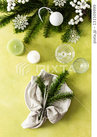 A top view of a Christmas table setting featuring an empty plate with napkin and cutlery, festive spruse and white tree decorations on a bright green background 131509999