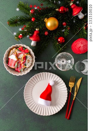 A Christmas table setting featuring an empty plate, napkin and gingerbread ,cutlery and a candle on a deep green background. Spruse branches and red tree decorations create a festive atmosphere 131510010