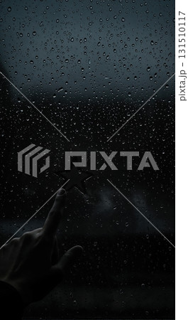 A person's hand touches a dark star shape on a window pane covered with numerous raindrops during a gloomy, atmospheric day. A person's hand touches a dark star shape on a window pane covered with numerous raindrops during a gloomy, atmospheric day. 131510117