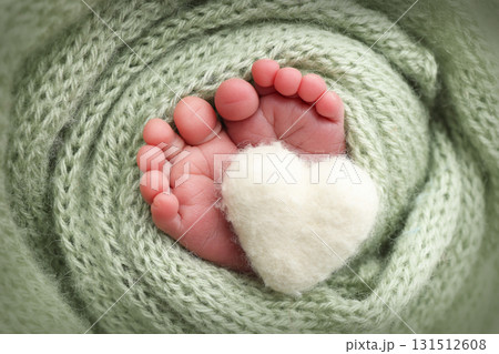 The tiny foot of a newborn baby. Soft feet of a new born in a olive green wool blanket. Close up of toes, heels and feet of a newborn. Knitted white heart in the legs of a baby. Macro photography. 131512608