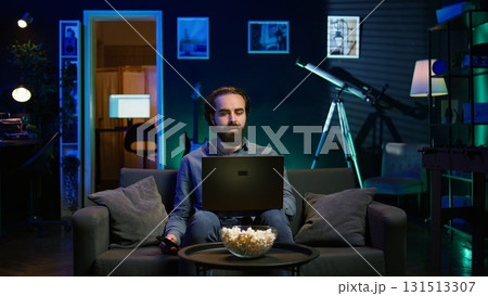 Man typing on laptop keyboard in neon lit apartment, sitting on couch and watching TV with bowl of popcorn ready. Teleworking person solving tasks on notebook with film playing in background, camera B 131513307