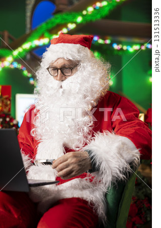 Santa Claus wearing red suit and glasses using laptop in North Pole location to monitor toy shipments worldwide. Elderly man dressed as xmas character follows gift deliveries on notebook 131513336